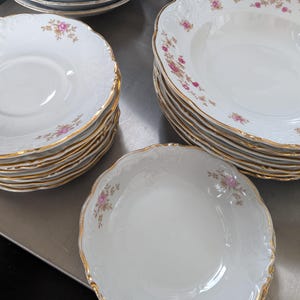 May include: A collection of white porcelain dishes with a gold rim and floral accents. The set includes various sizes of plates and bowls, stacked neatly. The floral design features pink and gold details, adding a touch of elegance to the tableware.