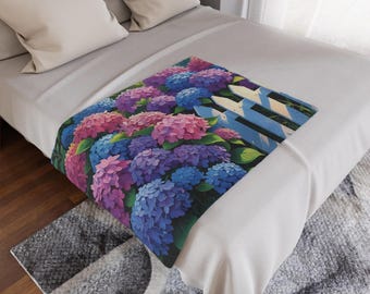 Op de afbeelding: Een sprei met een levendig bloemmotief van roze, paarse en blauwe hortensia's met een wit en blauw hek. De sprei ligt op een bed met witte lakens en kussens en een grijs tapijt.
