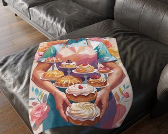 Puede incluir: Una manta suave con una ilustración en acuarela de una persona sosteniendo platos de pasteles. El diseño incluye varios pasteles y elementos florales en tonos de rosa, naranja y azul. La manta se muestra en un sofá de cuero negro.