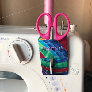 May include: A colourful fabric scissor holder with the word "Momma" embroidered on it. A pair of pink scissors is attached to the holder.