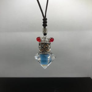 Può includere: Un ciondolo di cristallo a forma di diamante con una parte superiore in filigrana color argento e accenti di perline rosse. Il ciondolo è appeso a un cordino marrone, creando un accessorio unico. Il cristallo ha una tonalità azzurra.