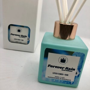 May include: A light blue glass bottle with a gold lid and white reeds. The bottle has a label that says "Forever Rain Reed Diffuser" and "Lotus Flower + Rain". The bottle is 100 ml or 3.3 oz and is hand-poured in Australia.