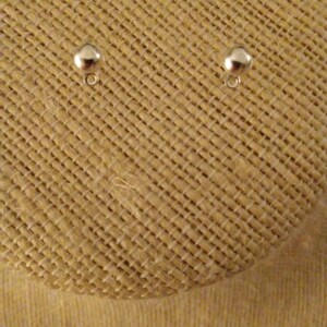 May include: Two silver-toned earring posts with round tops, displayed on a beige fabric surface.