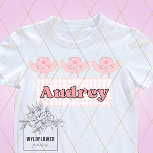 May include: White t-shirt featuring three pink cowboy hats and a pink and white gingham banner with the name "Audrey" printed in pink.
