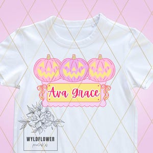 Può includere: Maglietta bianca con un design rosa e giallo di tre zucche di jack-o'-lantern con il testo "Ava Grace" su uno stendardo rosa.