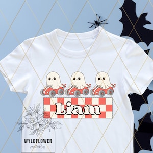 Op de afbeelding: Wit T-shirt met een rood-wit-zwart geruit ontwerp. Drie witte geesten rijden in rode raceauto's. De tekst "Liam" is op het geruite ontwerp gedrukt.
