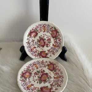 May include: Two white ceramic plates with a floral design in shades of red, pink, yellow, and green. The plates have a scalloped edge and are displayed on a black stand. The plates are on a white fur-like surface.