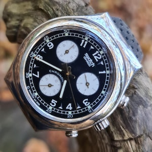 May include: A silver-tone Swatch Chrono wristwatch with a black dial, three white sub-dials, and white Arabic numerals. The watch face features the text Swatch Swiss and Chrono, with Swatch AG 1995 printed near the bottom edge of the dial.