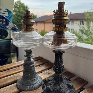 May include: Two oil lamps with clear glass reservoirs and brass burner assemblies. The left lamp features a grey metal base with embossed figures, while the right lamp has a dark grey, ornate metal base with floral patterns and a visible wick.