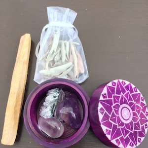 May include: A purple wooden box with a carved floral design on the lid. The box is open and contains a small bag of white sage, a purple amethyst crystal, and a clear quartz crystal. A wooden stick of incense is also included.