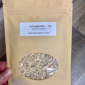 May include: A brown paper bag with a white label that reads "Ashwagandha 56g Withania somnifera Gratitudeandmore.com". The bag contains dried herbs.