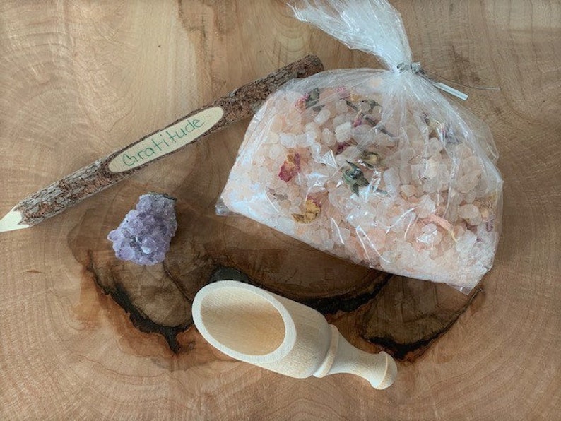 May include: A wooden stick with the word "Gratitude" written on it, a small amethyst crystal, a wooden scoop, and a bag of pink Himalayan salt with dried flowers.