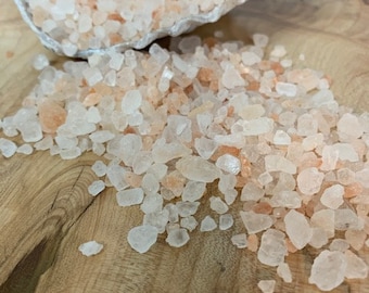 May include: Pink Himalayan salt crystals in a seashell on a wooden surface. The salt crystals are small and irregular in shape.