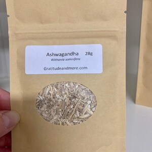 May include: A brown paper bag with a white label that reads "Ashwagandha 28g Withania somnifera Gratitudeandmore.com". The bag has a clear window showing dried herbs inside.