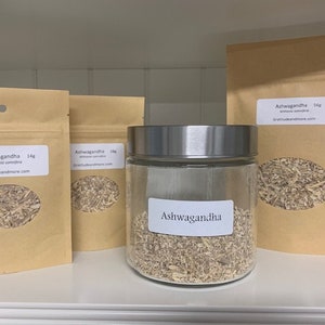 May include: Three brown paper bags with white labels that say "Ashwagandha" and a glass jar with a silver lid filled with dried herbs. The labels on the bags say "Ashwagandha" and "Withania somnifera" and include the website "gratitudeandmore.com".
