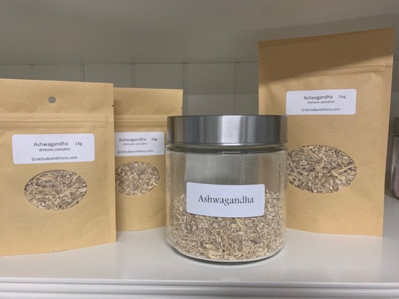 May include: Three brown paper bags with white labels that say "Ashwagandha" and a glass jar with a silver lid filled with dried herbs. The labels on the bags say "Ashwagandha" and "Withania somnifera" and include the website "gratitudeandmore.com".