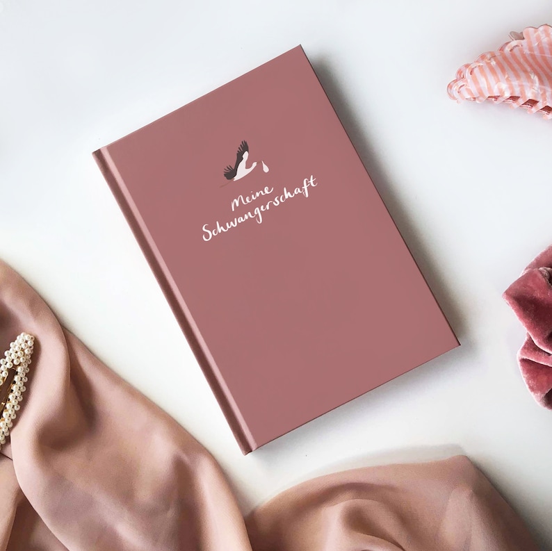 May include: A dusty rose colored journal with the text "Meine Schwangerschaft" and a white stork illustration.