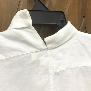 Puede incluir: Camisa blanca de lino con cuello y cierre de botones. La camisa est&aacute; colgada en una percha negra.