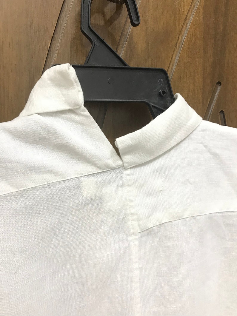 Puede incluir: Camisa blanca de lino con cuello y cierre de botones. La camisa est&aacute; colgada en una percha negra.
