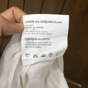Puede incluir: Etiqueta de cuidado blanca con texto negro que dice "COMME des GAR&Ccedil;ONS Co., Ltd. JM-0041 MADE IN JAPAN 100% LINEN HAND WASH. DO NOT BLEACH. DO NOT IRON. DRY CLEAN. LINE DRY. FABRIQUE AU JAPON 100% LIN LAVAGE MAINS. JAVEL INTERDIT. NE PAS REPASSER. NETTOYAGE A SEC. SECHAGE A L'OMBRE." La etiqueta tiene un fondo blanco con texto negro y s&iacute;mbolos para las instrucciones de lavado.