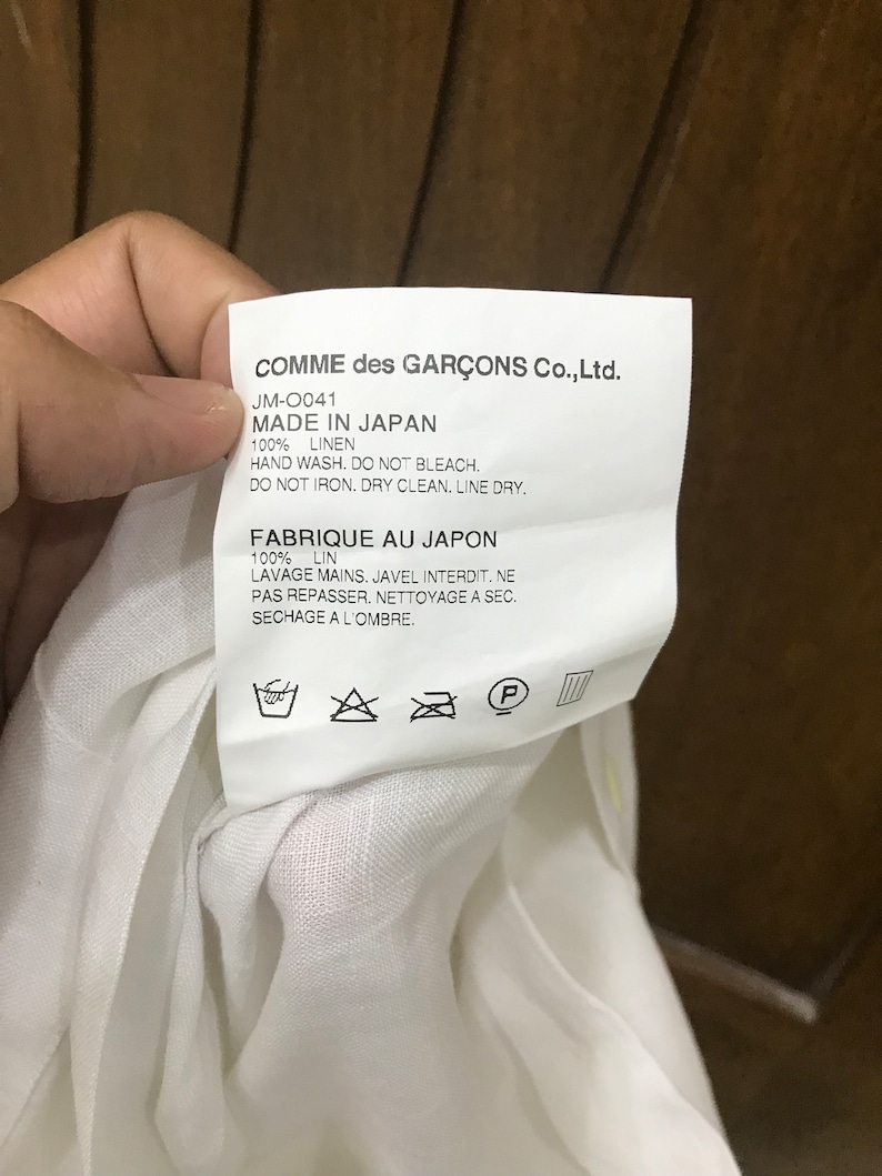 Puede incluir: Etiqueta de cuidado blanca con texto negro que dice "COMME des GAR&Ccedil;ONS Co., Ltd. JM-0041 MADE IN JAPAN 100% LINEN HAND WASH. DO NOT BLEACH. DO NOT IRON. DRY CLEAN. LINE DRY. FABRIQUE AU JAPON 100% LIN LAVAGE MAINS. JAVEL INTERDIT. NE PAS REPASSER. NETTOYAGE A SEC. SECHAGE A L'OMBRE." La etiqueta tiene un fondo blanco con texto negro y s&iacute;mbolos para las instrucciones de lavado.