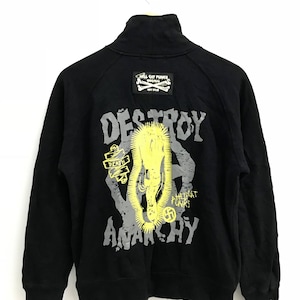 Könnte beinhalten: Schwarzer Reißverschluss-Sweatshirt mit einem grafischen Druck einer Hand mit einem Heiligenschein und den Worten "Destroy Anarchy" in Gelb und Grau. Der Text "Hell Cat Punx Original" ist auf einem kleinen Patch auf der Rückseite des Sweatshirts aufgedruckt.