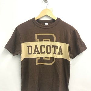 May include: Brown short-sleeved t-shirt with a large tan letter "D" and the word "DACOTA" printed on the front.