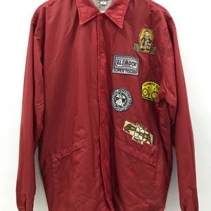 Può includere: Giacca di nylon rossa con più patch ricamate. I patch includono "Elsmock Super Tough", "Clanky's Cycles" e "Strong Parts".