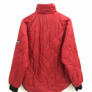 May include: A red quilted jacket with a black zipper and black ribbed cuffs. The jacket has a small logo patch on the left sleeve that says "Bear.