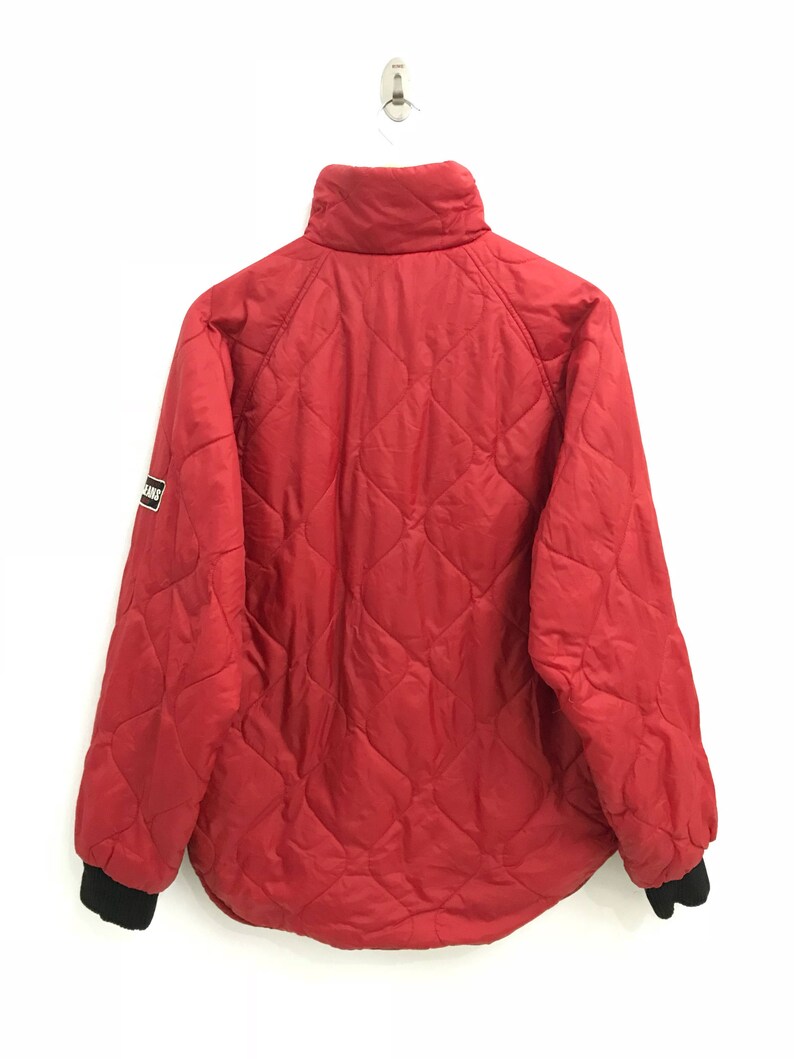 May include: A red quilted jacket with a black zipper and black ribbed cuffs. The jacket has a small logo patch on the left sleeve that says "Bear.