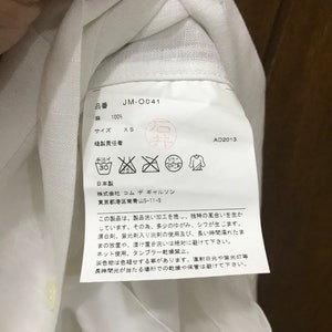 Puede incluir: Etiqueta de cuidado de la ropa blanca con texto japon&eacute;s, incluyendo el nombre de la marca "Commes des Garcons" y el n&uacute;mero de producto "JM-0041". La etiqueta tambi&eacute;n incluye instrucciones de cuidado, talla y contenido de la tela.