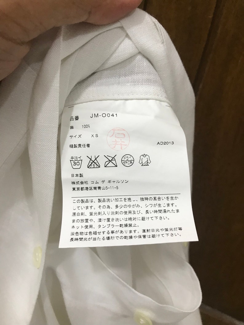 Puede incluir: Etiqueta de cuidado de la ropa blanca con texto japon&eacute;s, incluyendo el nombre de la marca "Commes des Garcons" y el n&uacute;mero de producto "JM-0041". La etiqueta tambi&eacute;n incluye instrucciones de cuidado, talla y contenido de la tela.