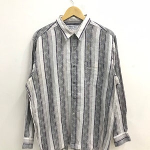 Puede incluir: Una camisa de manga larga con botones y un patrón de rayas blancas y grises. La camisa tiene un pequeño bolsillo en el pecho izquierdo y una etiqueta que dice "Roger Waters".