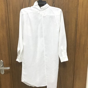 Puede incluir: Camisa blanca de manga larga con cuello alto y dobladillo asim&eacute;trico. La camisa est&aacute; colgada de una percha contra una puerta de madera.