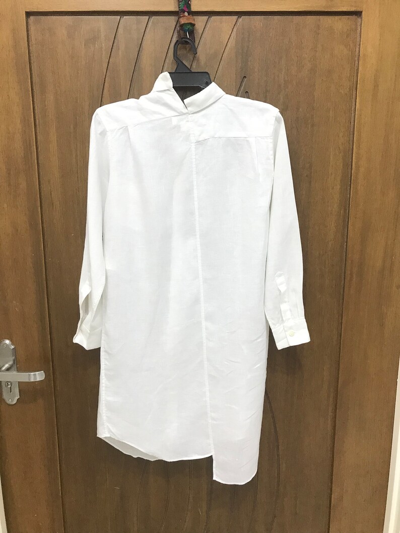 Puede incluir: Camisa blanca de manga larga con cuello alto y dobladillo asim&eacute;trico. La camisa est&aacute; colgada de una percha contra una puerta de madera.