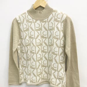 May include: A beige turtleneck sweater with a white geometric pattern. The pattern is made up of repeating letters.