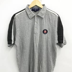Op de afbeelding: Een grijs poloshirt met korte mouwen met zwarte en witte strepen op de mouwen. Het shirt heeft een klein logo op de linkerborst met de tekst "Polo Sport" met een rode "P" in een cirkel.