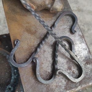 Op de afbeelding: Vier handgesmede metalen haken met een gedraaid ontwerp. De haken liggen op een metalen oppervlak.