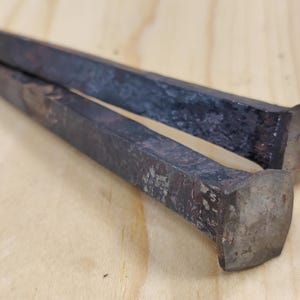 Puede incluir: Dos puntas metálicas antiguas y oscuras, con cabezas cuadradas y extremos puntiagudos, sobre una superficie de madera clara. Las puntas probablemente se utilizan para proyectos de construcción o carpintería.