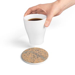 May include: A white ceramic mug filled with dark liquid and a round cork coaster featuring a detailed, printed map design in shades of tan, brown, and blue, depicting a city layout with a winding river running through the center.