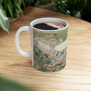 May include: White ceramic coffee mug with a detailed map design. The mug is filled with dark coffee and has a white handle. The map features a city with green hills and roads.