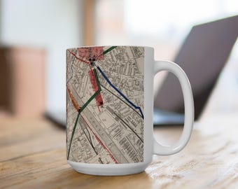 May include: A white ceramic mug featuring a detailed vintage map print. The map displays black and white street layouts with highlighted red, green, and blue lines. The mug has a cylindrical body and a curved handle on the right side.