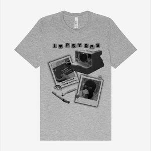 May include: A heather grey t-shirt with a black and white graphic design. The graphic features a Polaroid camera, a photo of a person wearing a black ski mask, and the text "I ♥ PSYOPS" and "MUSIC BOYFRIEND I'M YOUR HUM-HUM".