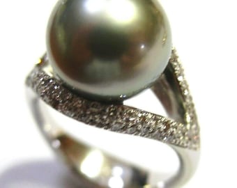 Pode incluir: Um anel de prata com uma grande pérola verde escura engastada em uma banda encrustada com diamantes.