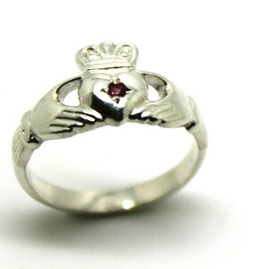 Peut inclure: Une bague Claddagh en argent avec un cœur rouge et une couronne sur le dessus. La bague présente deux mains jointes de chaque côté du cœur.