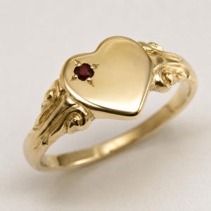 May include: A gold ring featuring a heart-shaped face with a small, round, deep red gemstone set into the upper left side. The band and the sides of the heart display raised, decorative scrollwork details that extend along the ring shank.