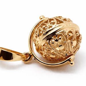 Könnte beinhalten: Ein goldener Anhänger mit einem filigranen Globus-Design. Der Anhänger hat eine detaillierte, durchbrochene Kugel, die in einem goldenen Rahmen gehalten wird, mit einer Schlaufe für eine Kette. Das Gesamtdesign ist aufwendig und elegant.