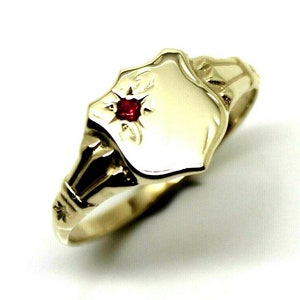 May include: A gold signet ring with a red gemstone set in the centre of the top. The ring has a shield-shaped top and a detailed band.