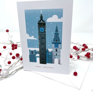 May include: A Christmas card featuring a winter scene with two tall clock towers under a snowy sky. The card has the words "MERRY CHRISTMAS" in a red font. A white envelope is visible.