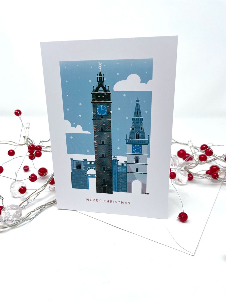 May include: A Christmas card featuring a winter scene with two tall clock towers under a snowy sky. The card has the words "MERRY CHRISTMAS" in a red font. A white envelope is visible.